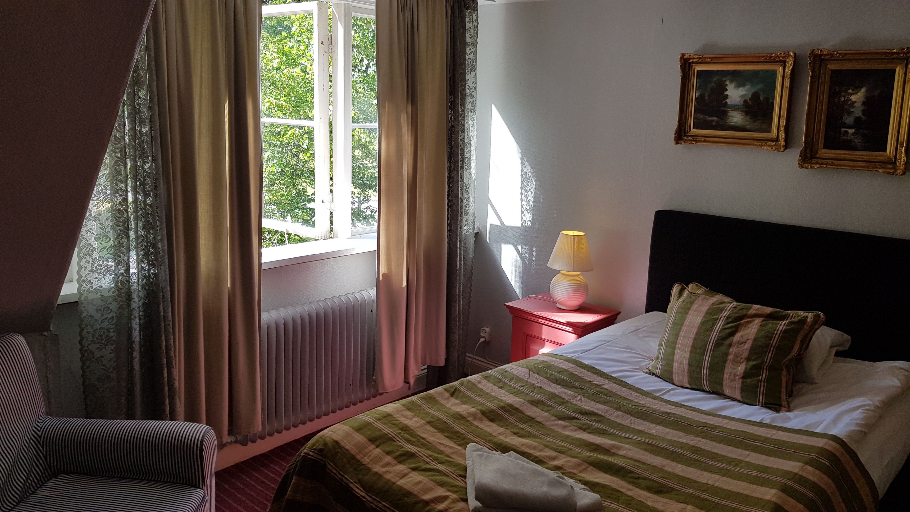 A hotel room with a bed, a chair, a nightstand with a lamp, and two framed pictures on the wall.