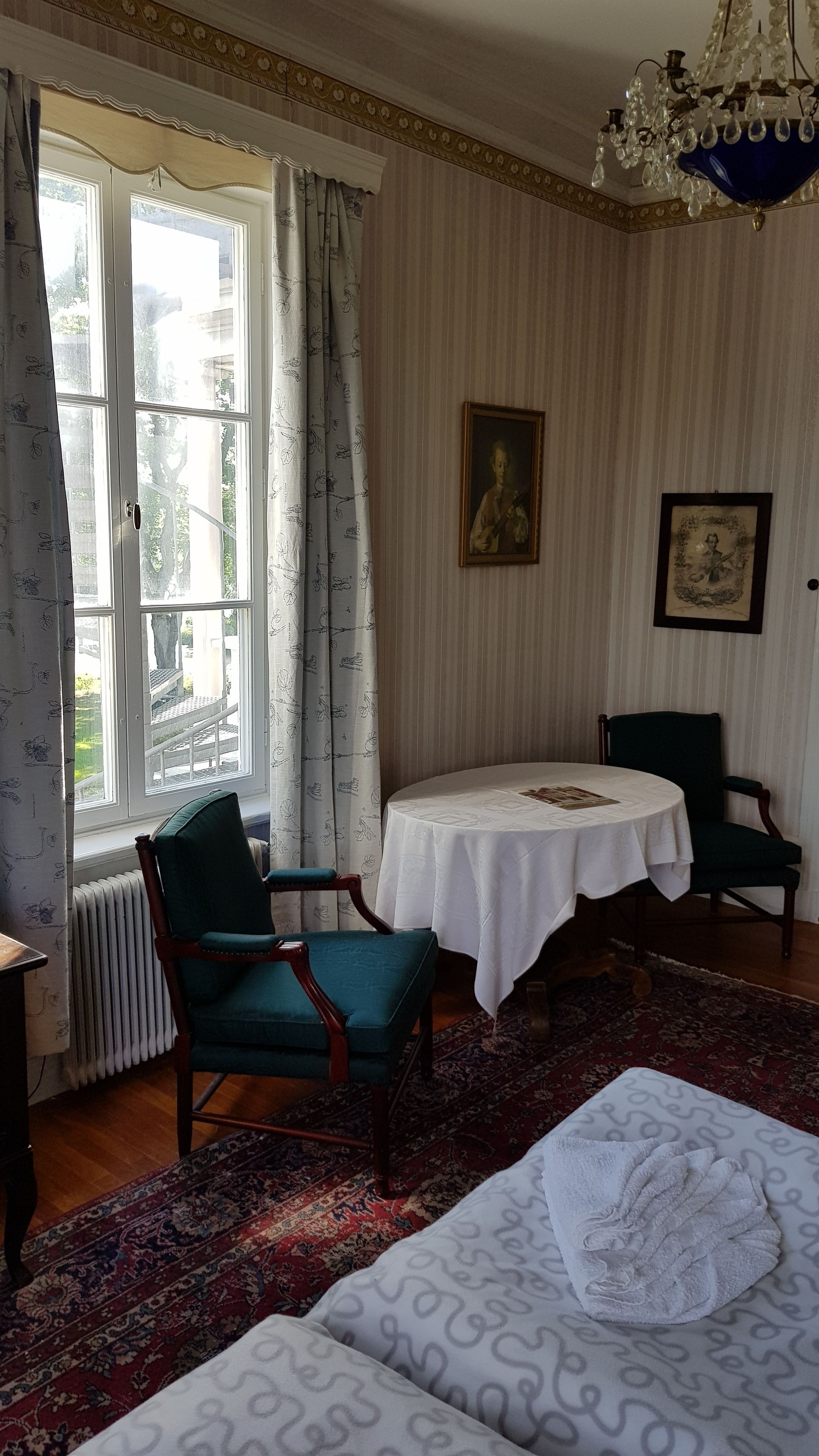 A room with a large window, a table with a white tablecloth, two chairs, a bed with a white cover, and a chandelier.