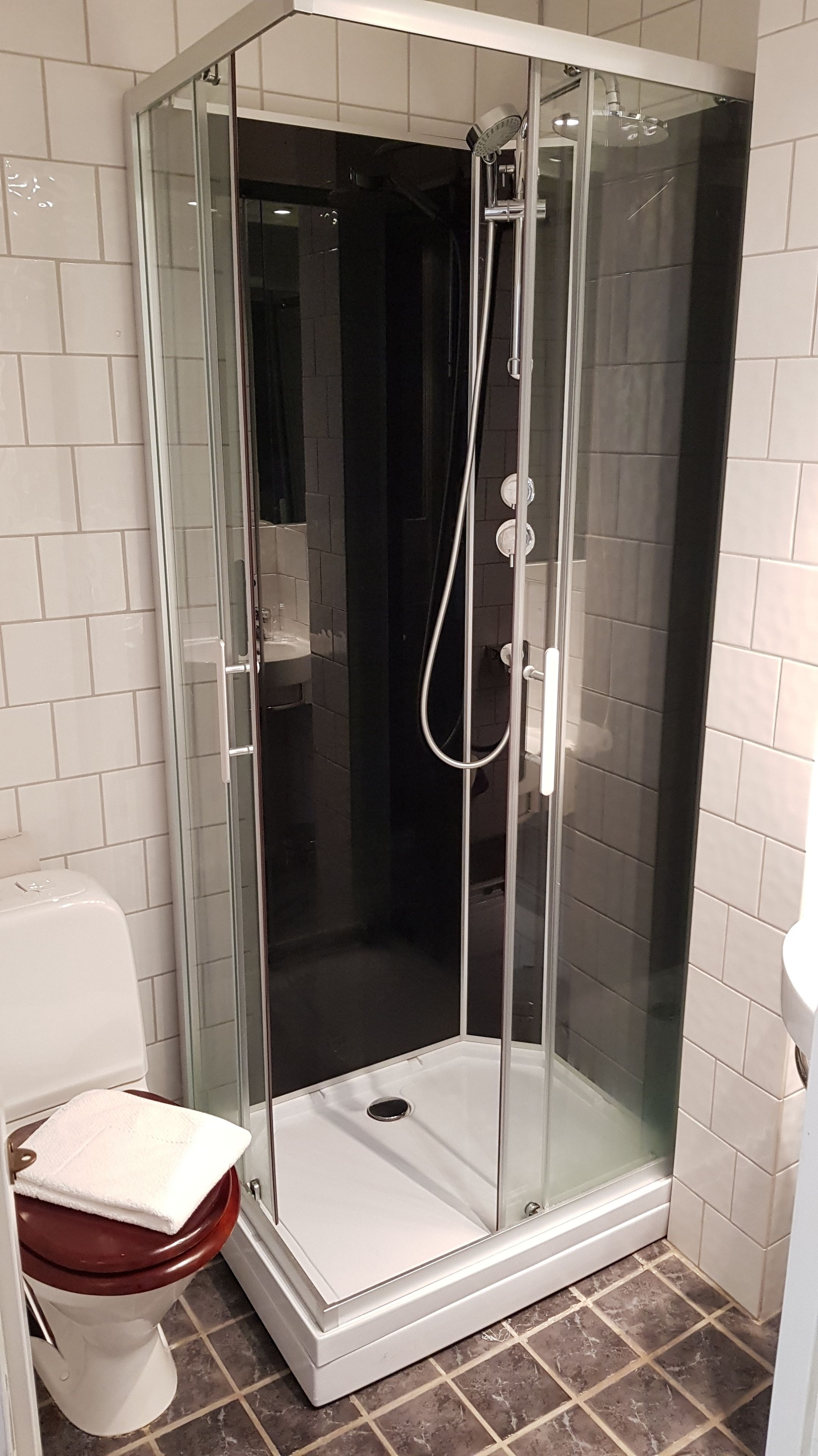 A glass-enclosed shower stall with a white tiled floor, adjacent to a toilet with a wooden seat.