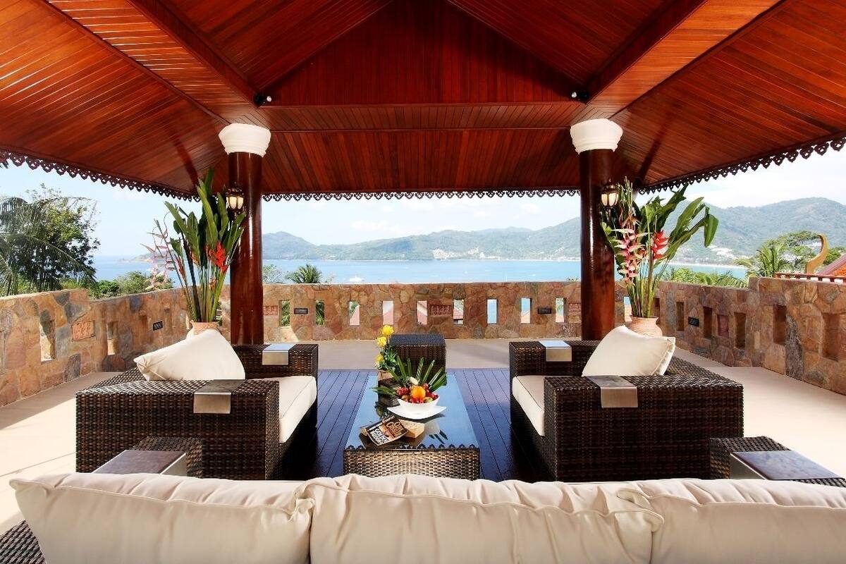 A covered outdoor seating area with wicker furniture and a table with fruit and flowers, overlooking a scenic view of the sea and mountains.