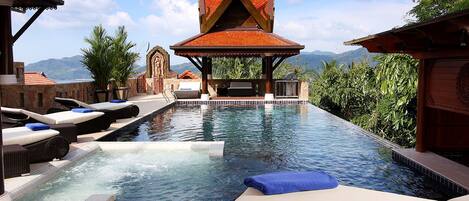 A pool area with lounge chairs and a covered seating area, surrounded by lush greenery and mountains in the background.