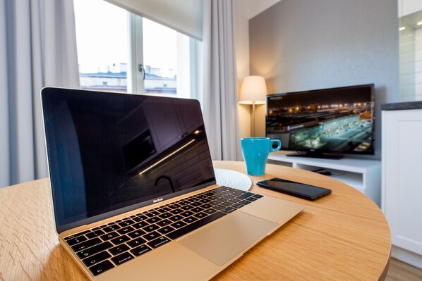 A laptop on a wooden table with a cityscape view, a blue mug, and a TV displaying a city scene. - Riverside Aparthotel (Kraków)