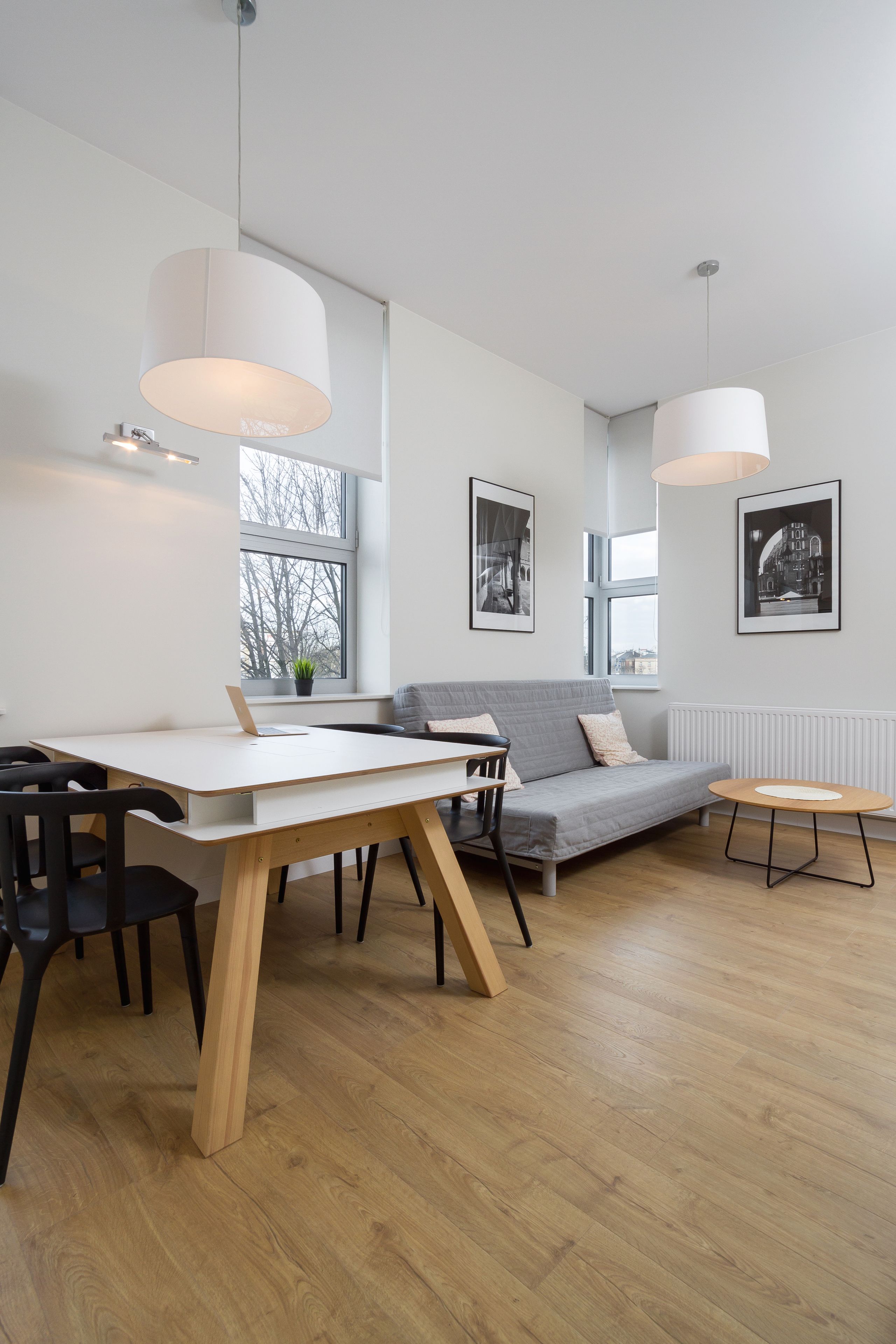 A modern living room with a white table, grey sofa, and a wooden floor. There are two pendant lights and a radiator.