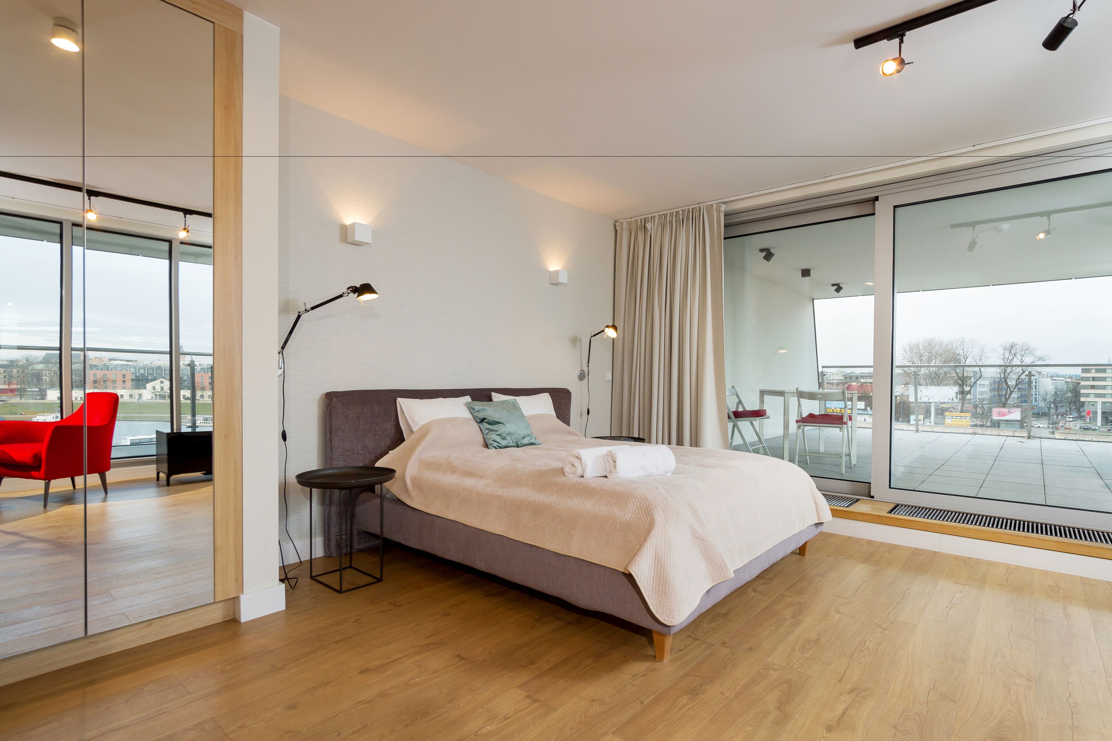 Un dormitorio moderno con una cama grande, una silla roja, una mesita pequeña y vistas a la ciudad a través de amplios ventanales.