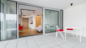 A modern hotel room with a sliding glass door leading to a bedroom. There is a white table with a red chair, a small white table, and a red folding chair.