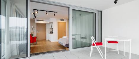 A modern hotel room with a sliding glass door leading to a bedroom. There is a white table with a red chair, a small white table, and a red folding chair.