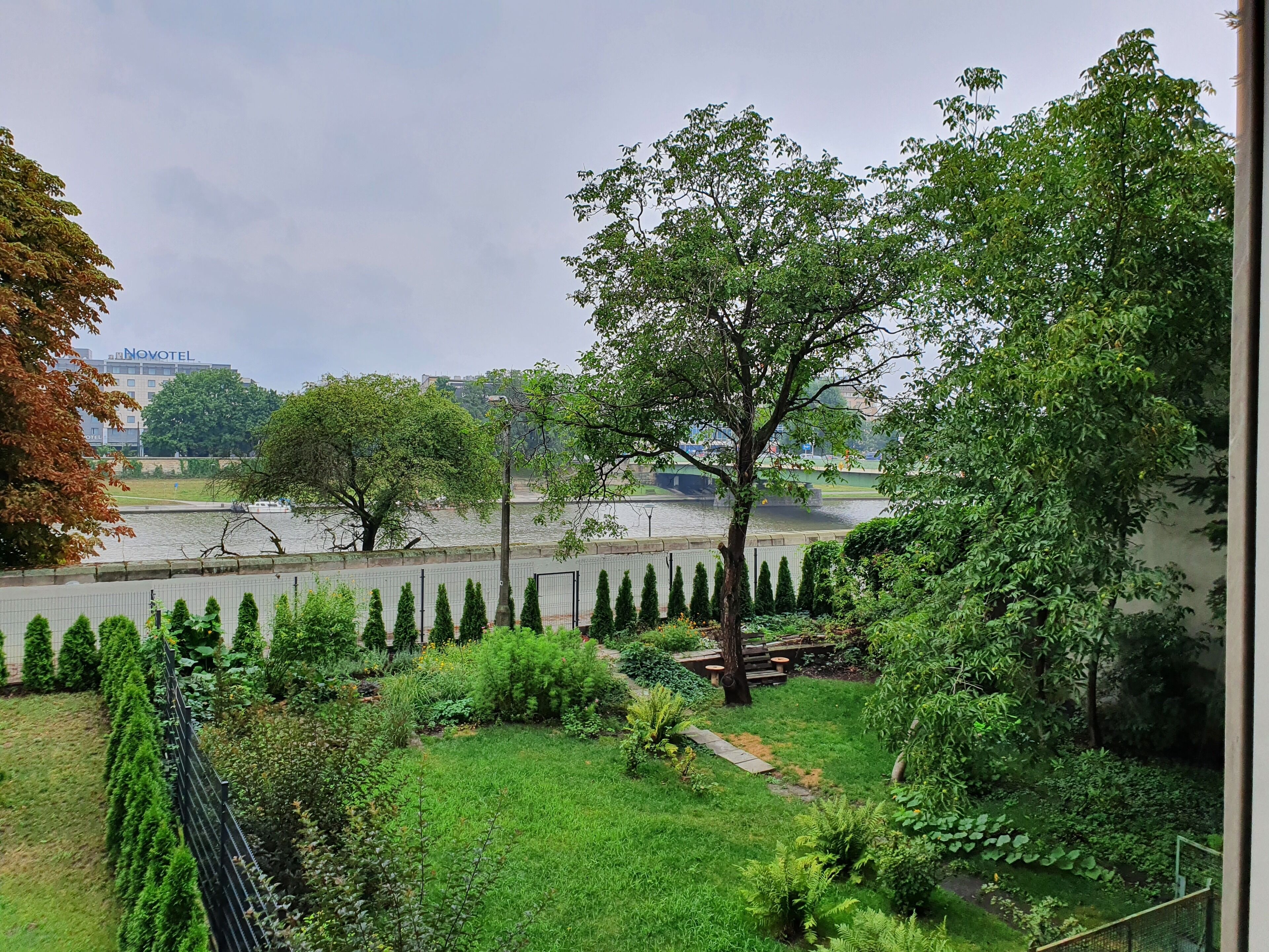 Un jardín con varios árboles y arbustos, una cerca y vistas de un río y edificios al fondo.