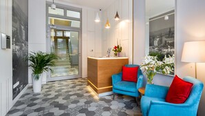 A modern reception area with a wooden desk, blue armchairs with red cushions, a potted plant, and a large mirror reflecting a cityscape.