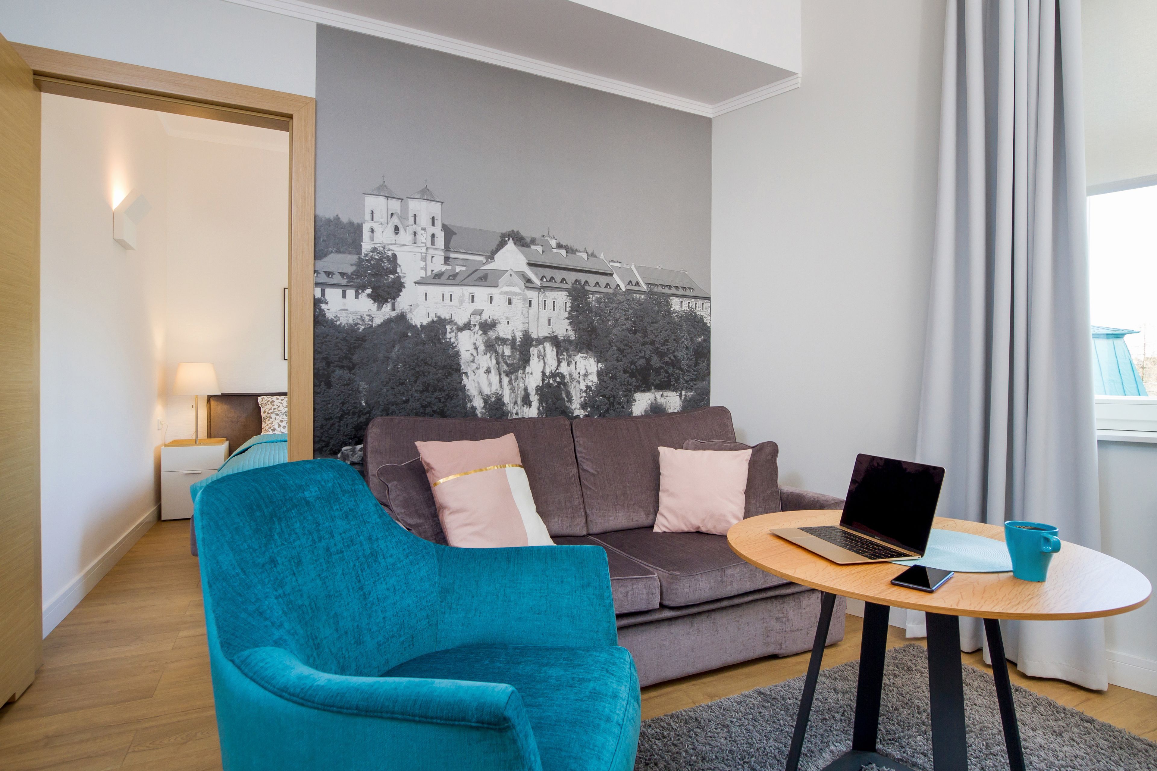 A modern room with a teal armchair, a grey sofa, a laptop on a wooden table, and a large black-and-white cityscape painting on the wall.