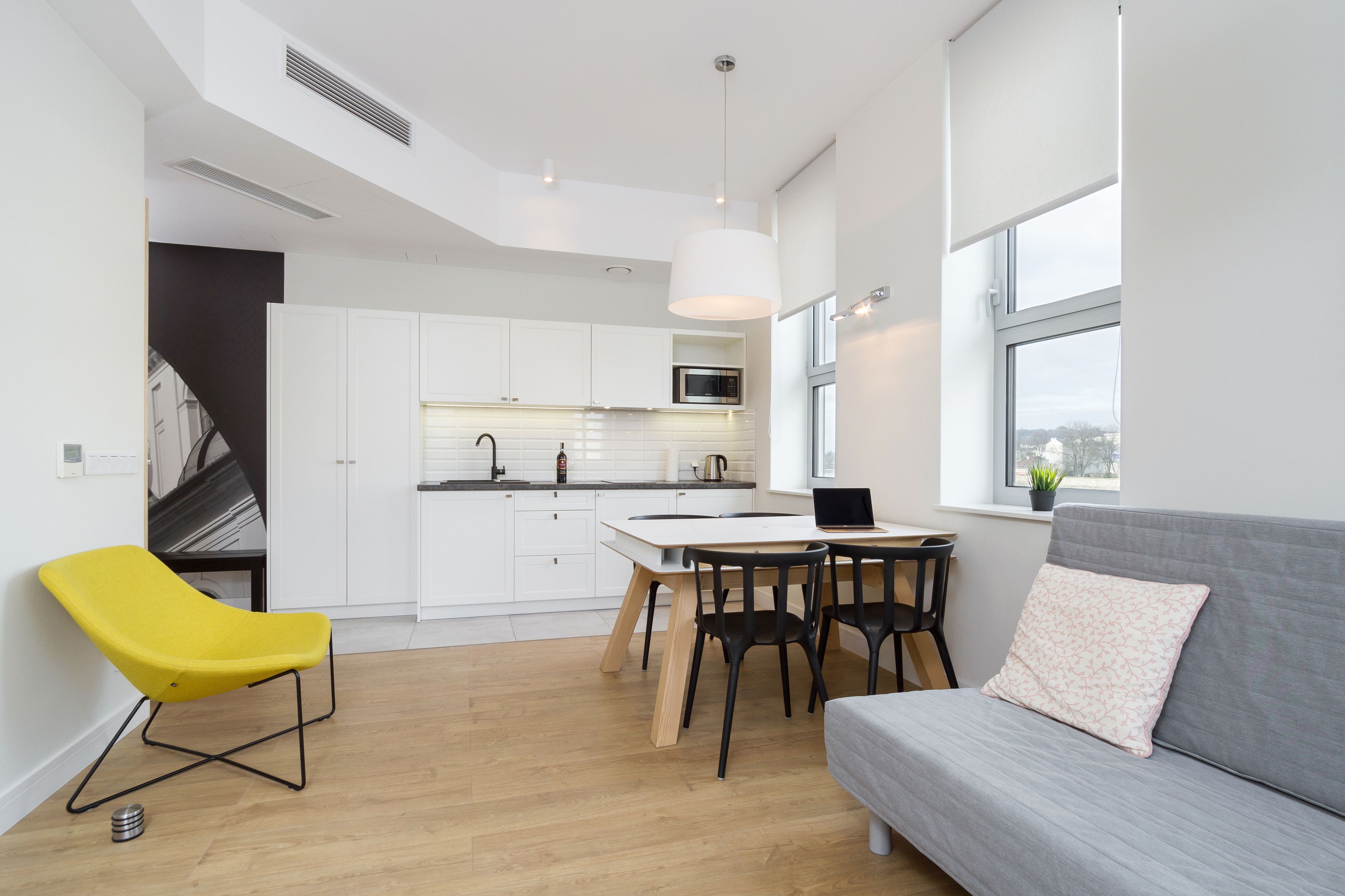 A modern kitchen with white cabinets, a dining table with black chairs, a yellow chair, and a grey sofa with a pillow.