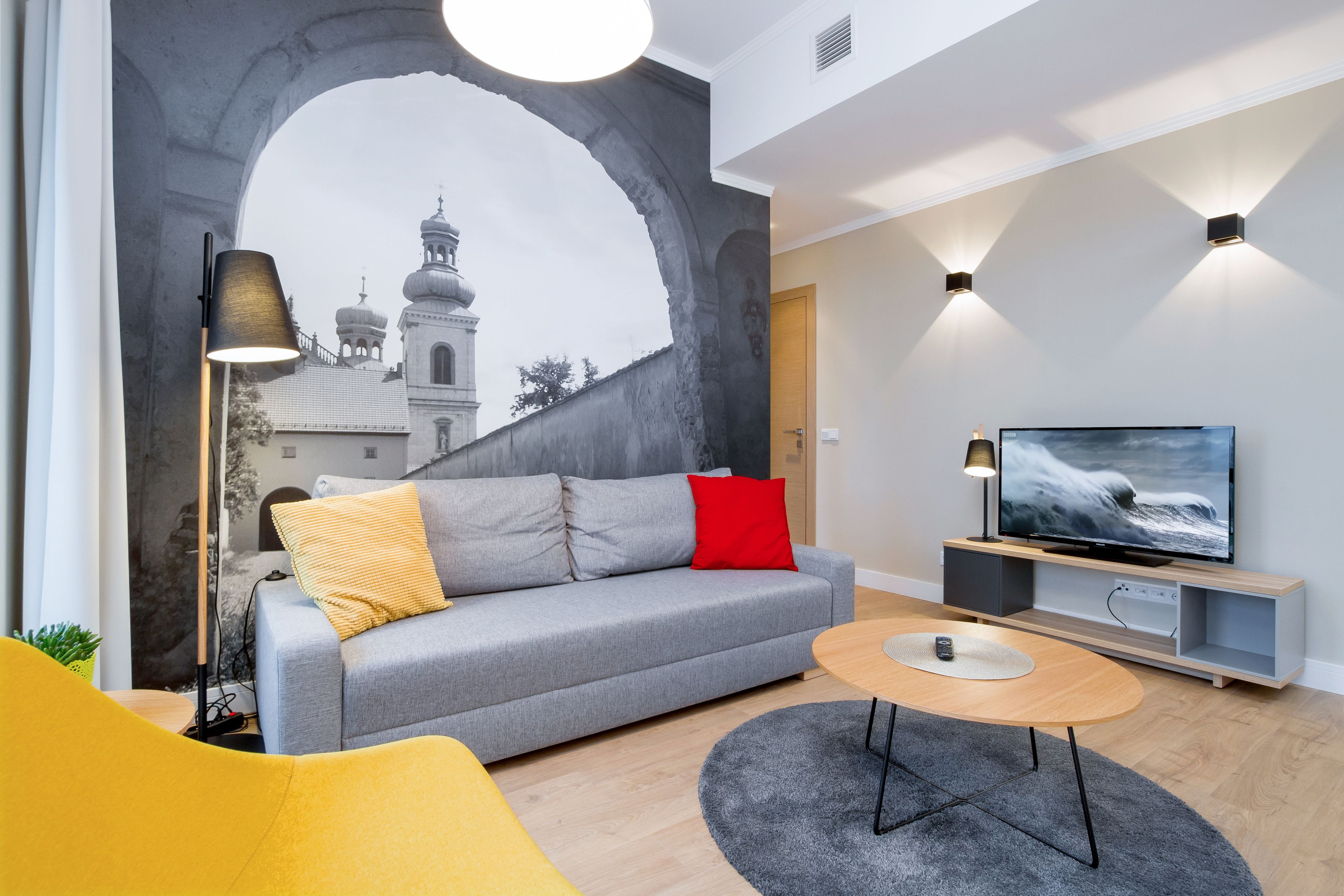 Un salon moderne avec un canapé gris, un fauteuil jaune et un téléviseur posé sur un meuble en bois.