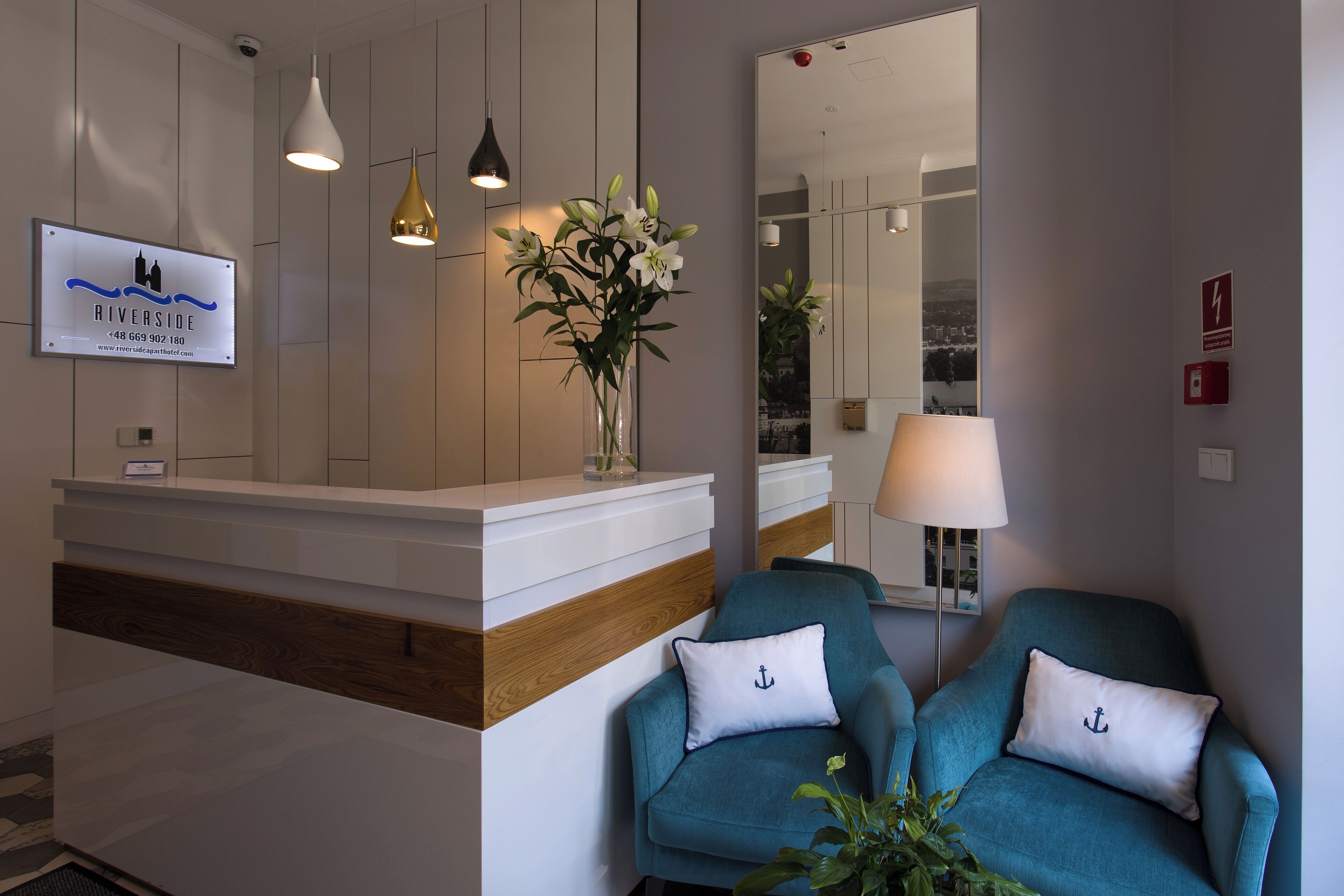 Un área de recepción con mostrador blanco, sillones azul turquesa, un jarrón con flores y un espejo que refleja la sala.