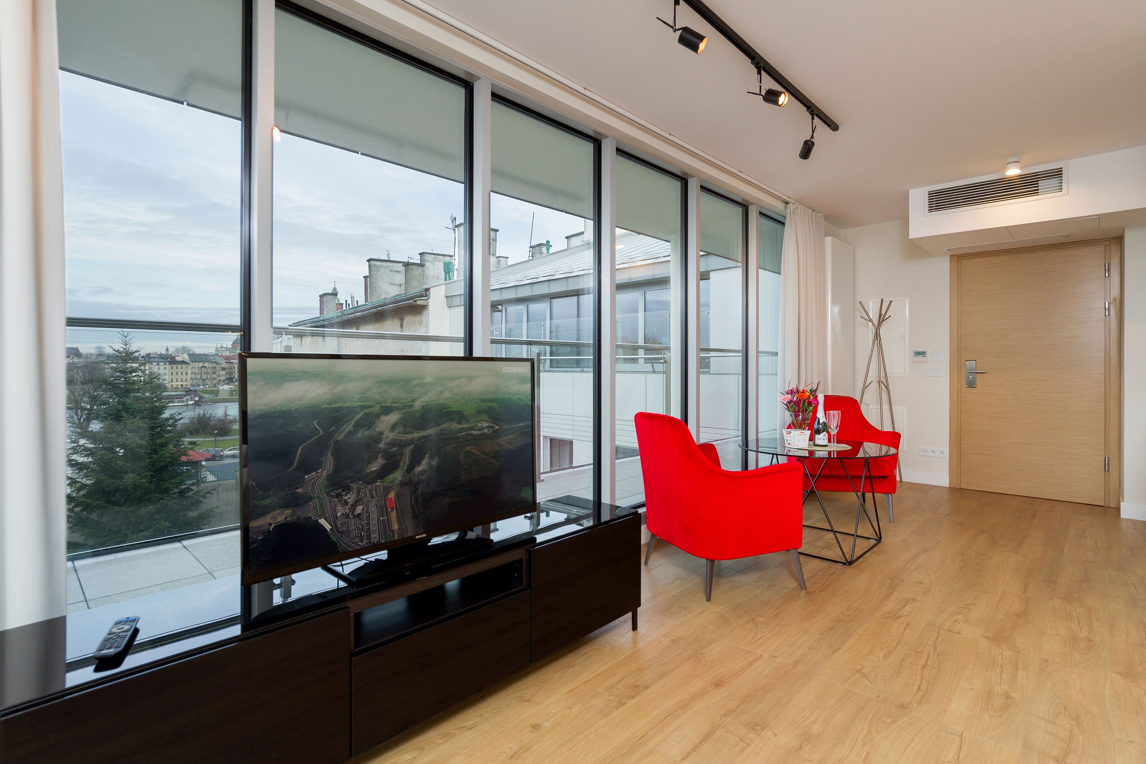 A modern living room with large windows, a flat-screen TV, a dining area with red chairs, and a wooden door.