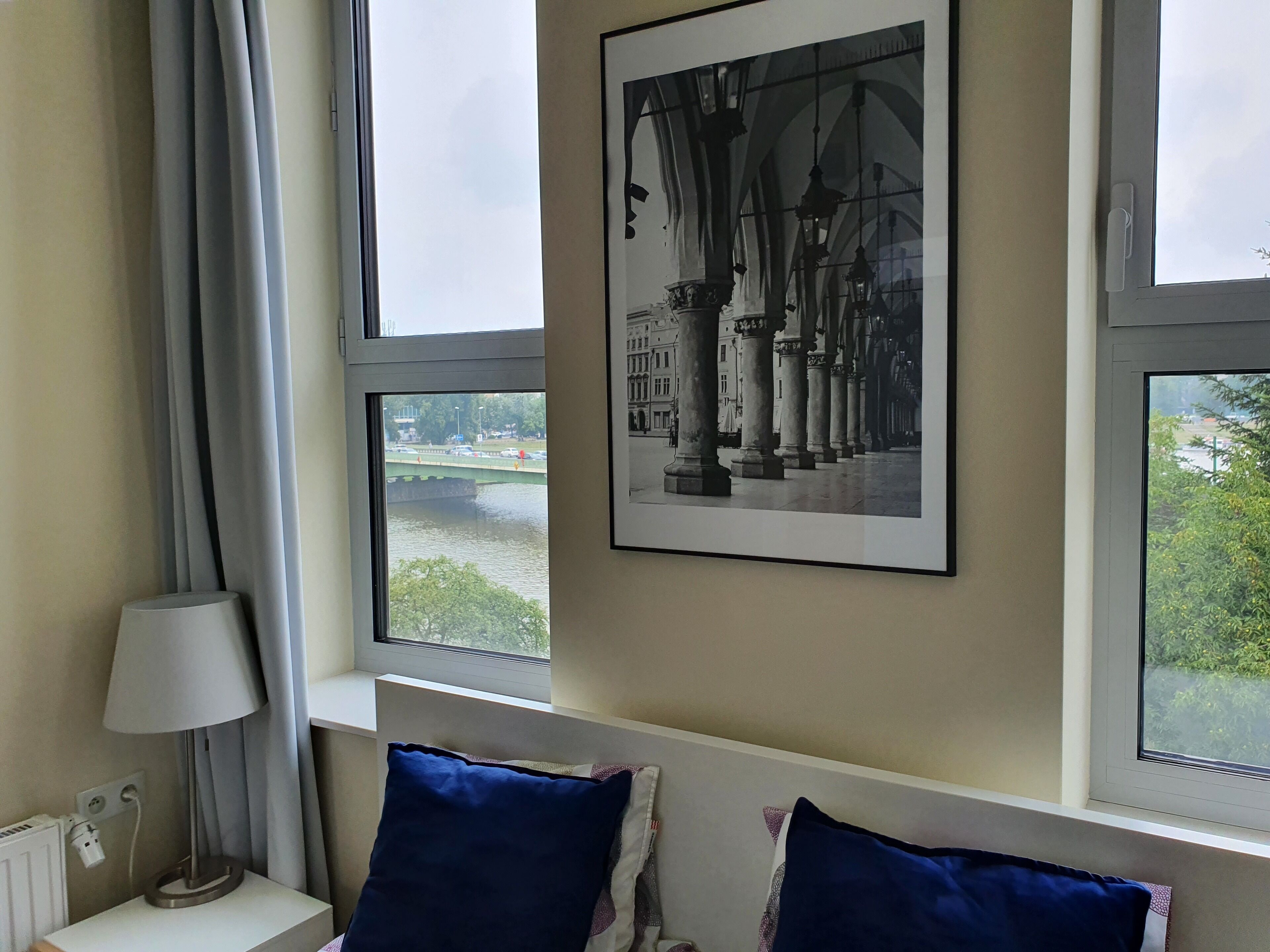 Ein Hotelzimmer mit einem großen Fenster, einem gerahmten Foto eines Gebäudes, einem Nachttisch mit Lampe und zwei blauen Kissen.