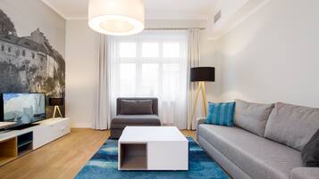 A modern living room with a flat-screen TV, a white coffee table, a grey sofa, a blue rug, and a large window with sheer curtains.