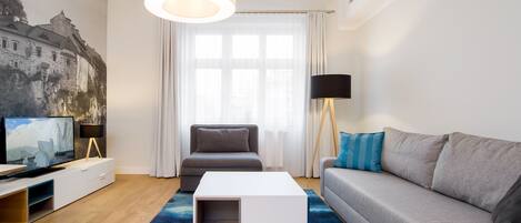 Una sala de estar moderna con un televisor de pantalla plana, una mesa de centro blanca, un sofá gris, una alfombra azul y un amplio ventanal con cortinas traslúcidas.