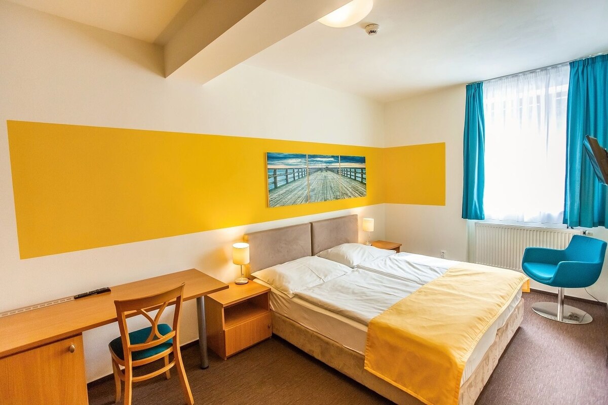 A hotel room with a bed, desk, chair, and a painting of a pier.