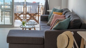 Un salón moderno con un sofá gris, cojines coloridos, una mesita redonda con vasos y una jarra, y vistas al mar a través de amplias ventanas.