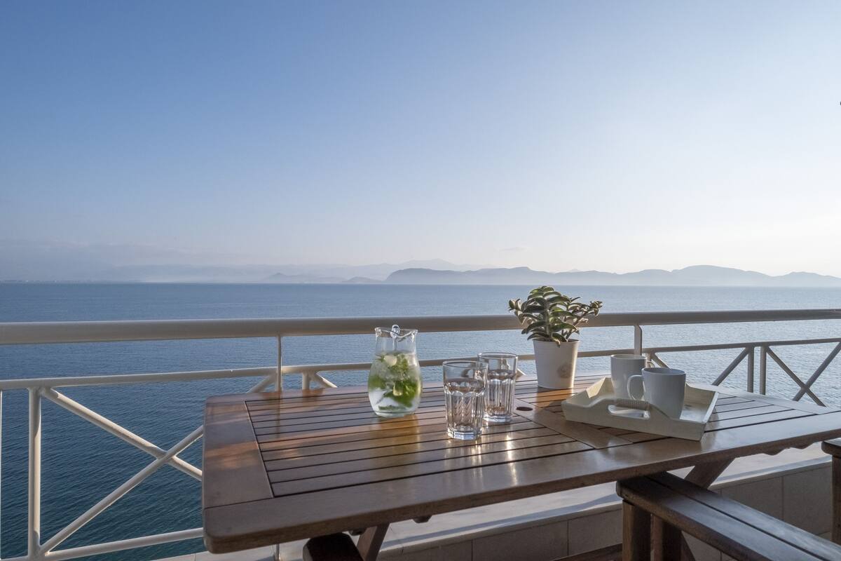 Una mesa de madera con una jarrón, vasos y una planta en maceta en un balcón con vista al mar.