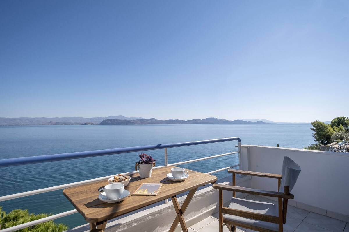 Una mesa de madera con tazas blancas y un menú en un balcón con vista a un mar tranquilo y montañas distantes.