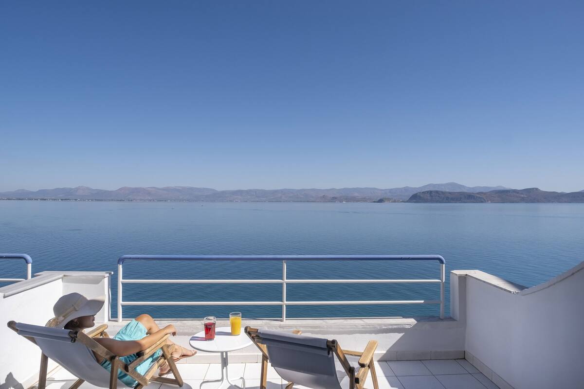 Un balcón con vistas a un mar tranquilo y montañas lejanas. Hay dos sillas y una mesita con una bebida.