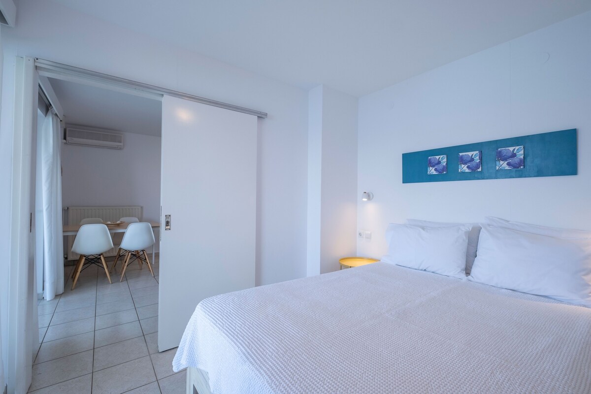 Un dormitorio con cama, cortinas blancas, una mesa con sillas y una obra de arte colgada en la pared.