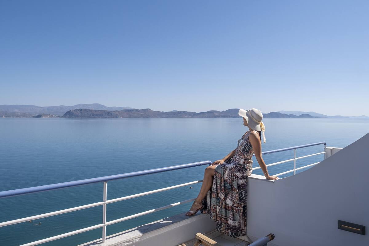 Una mujer sentada en un balcón con vista al mar tranquilo y montañas a lo lejos.