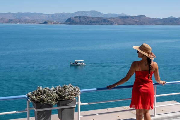 Une femme vêtue d’une robe rouge et coiffée d’un chapeau de paille se tient sur un balcon d’où l’on aperçoit une mer calme, avec des montagnes en arrière-plan.