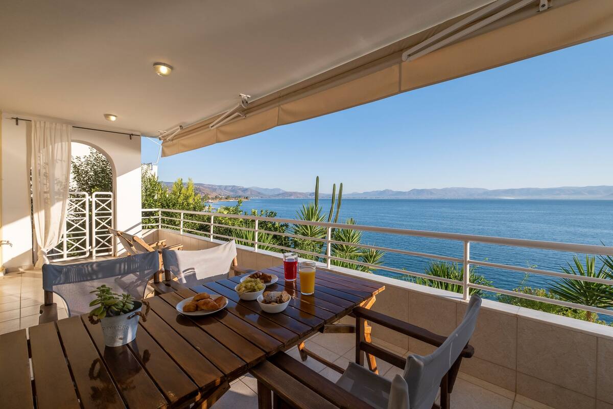 Mesa de madera con alimentos para el desayuno y una planta en maceta en un balcón con vistas al agua.