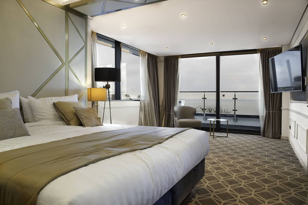 A hotel room with a large bed, a desk, a chair, a TV, and a balcony with a view.