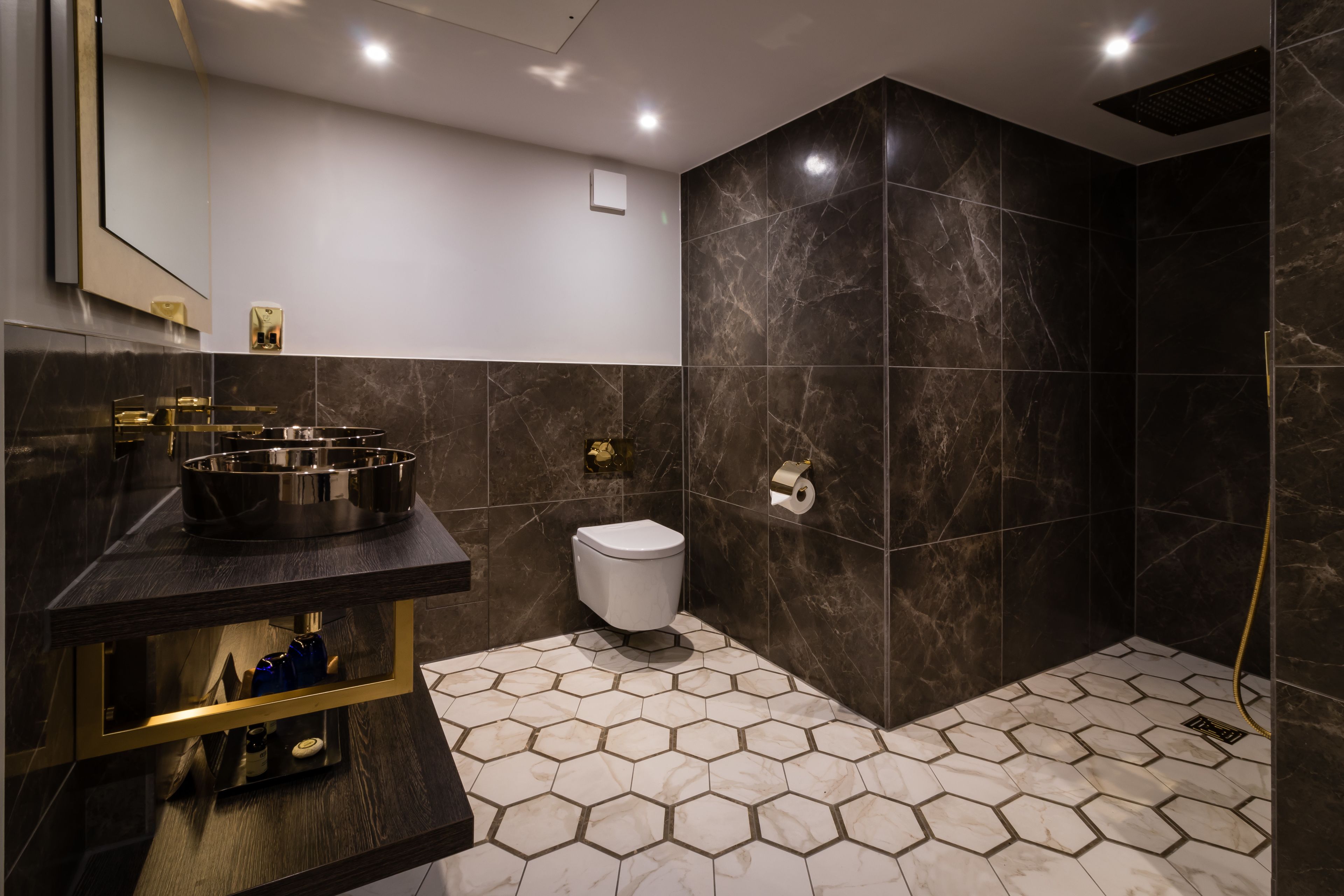 A modern bathroom with a hexagonal tile floor, a large mirror, a sink with a gold faucet, a toilet, and a shower area with dark tiles.
