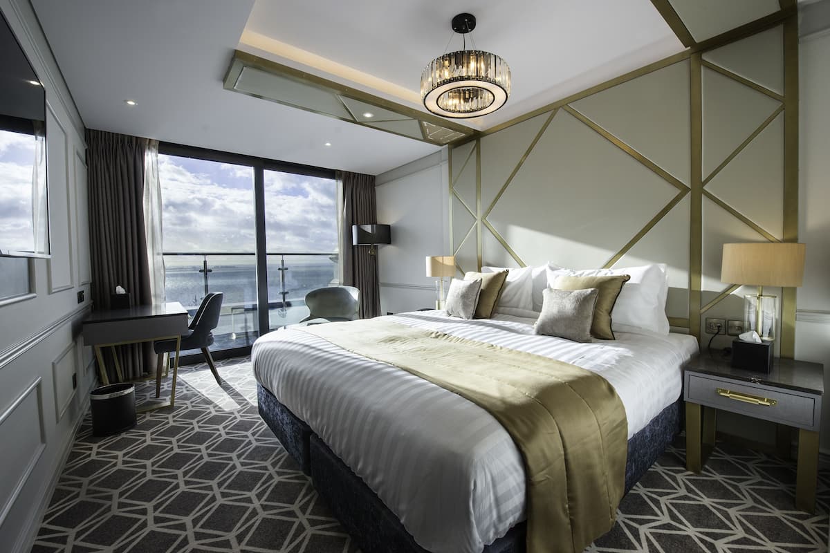 A modern hotel room with a large bed, a desk, and a chair. There is a view of the sky and clouds through the window.