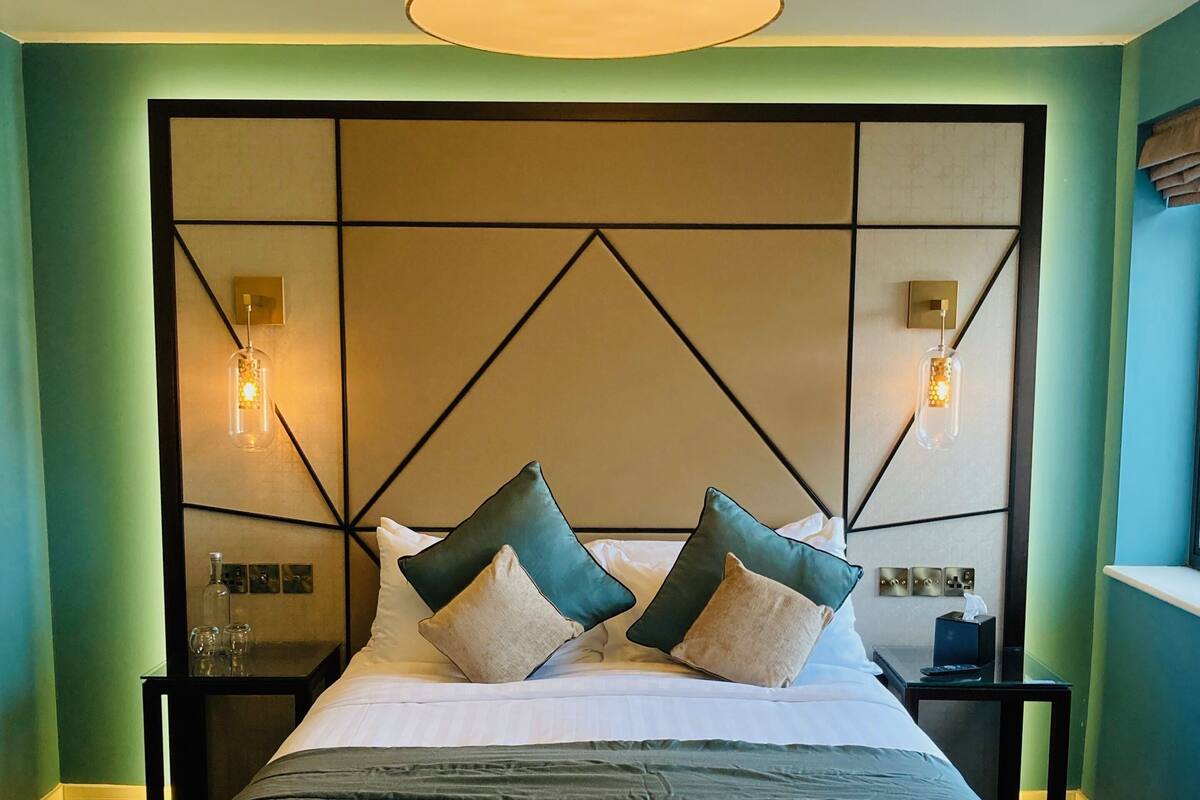 A hotel room with a large bed, a geometric headboard, bedside tables with lamps, and a hanging light fixture.