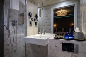 A modern bathroom with a glass-enclosed shower, a white sink with two faucets, and a mirror above the sink. There are bottles of toiletries on the sink's edge. - Seven Hotel (Southend-on-Sea)