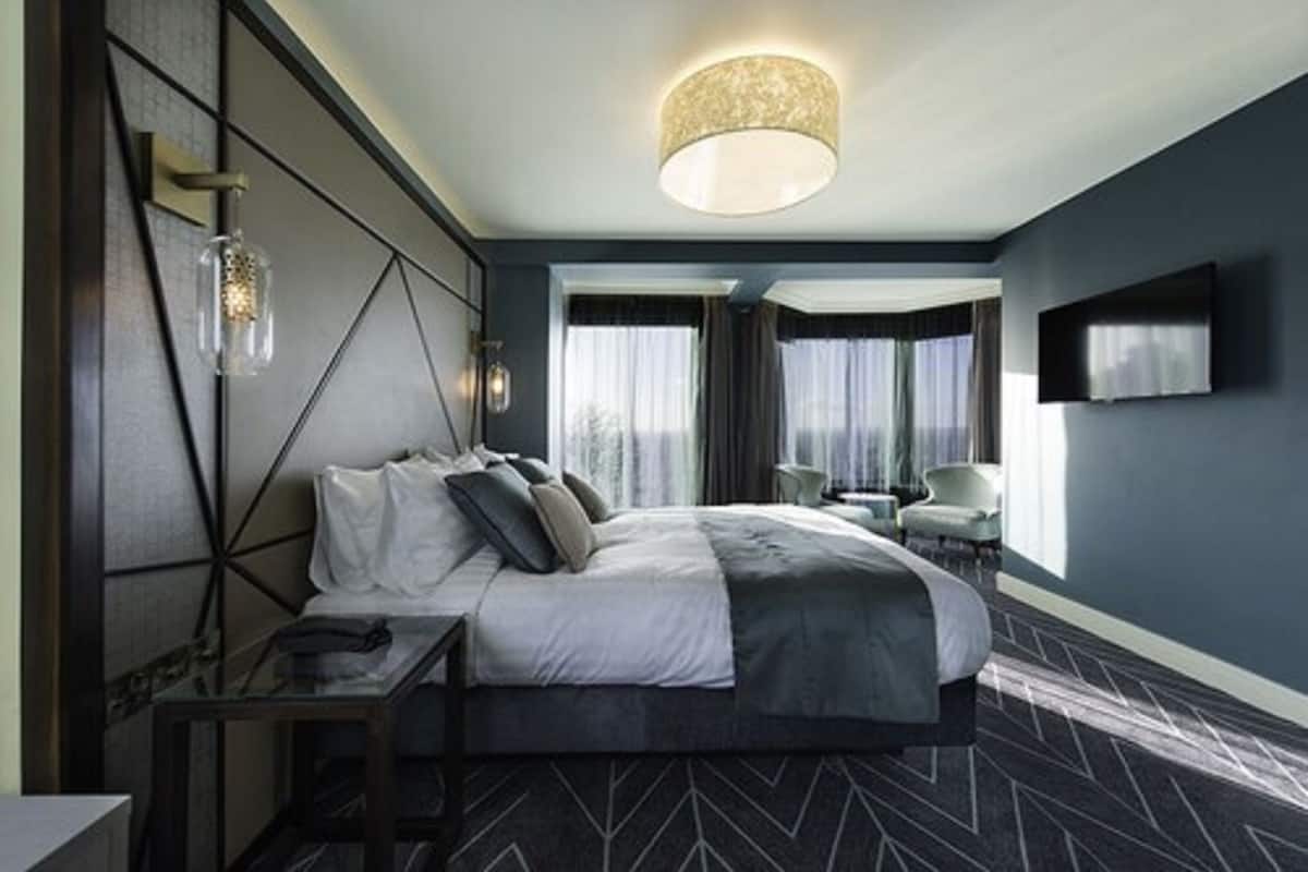 A modern hotel room with a large bed, a bedside table, a TV mounted on the wall, and a chandelier.