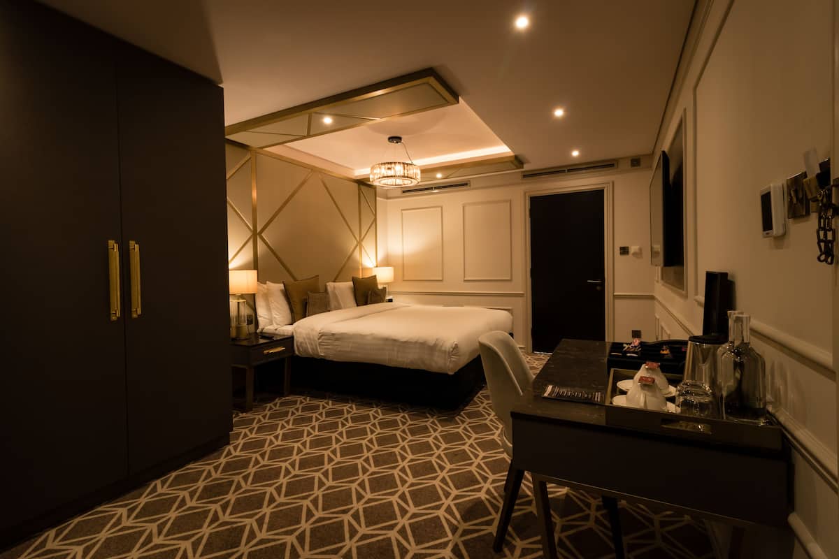 A modern hotel room with a geometric patterned carpet, a large bed, a desk with a chair, a nightstand, and a chandelier.