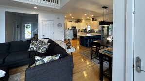A spacious living room with a dark-colored sofa and a dining area with a table and chairs. A kitchen with white cabinets and a large island is visible in the background.