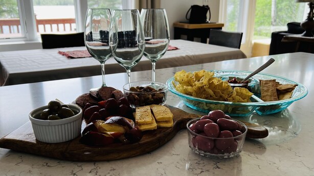 A dining table with a variety of foods and drinks, including olives, cured meats, cheese, and chips, set against a backdrop of large windows with a view of trees and a body of water.