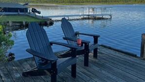 A wooden dock with two Adirondack chairs and a small table, overlooking a calm lake with a dock extending into the water.