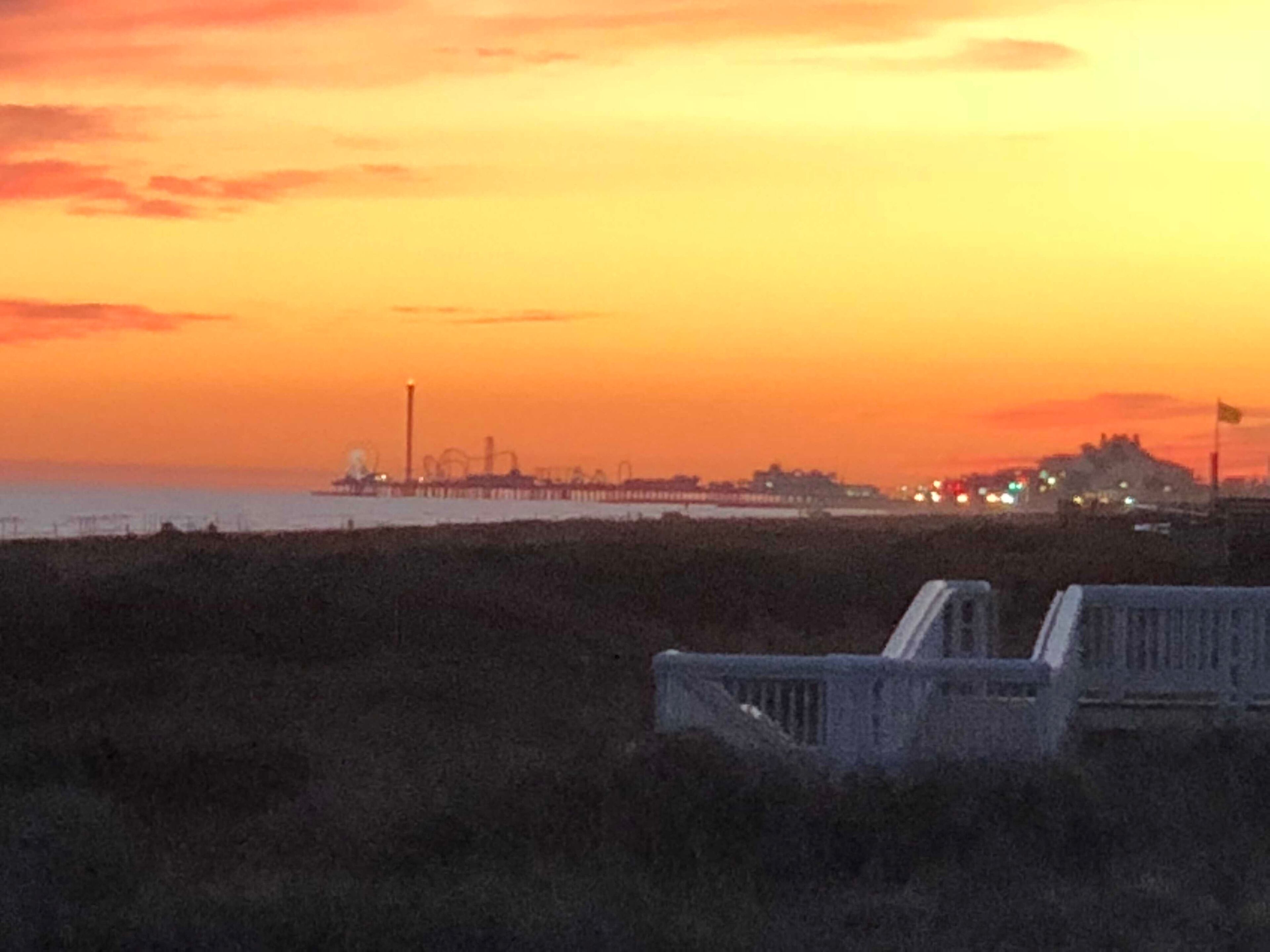 Een kustzonsondergang met een pier en gebouwen in de verte.