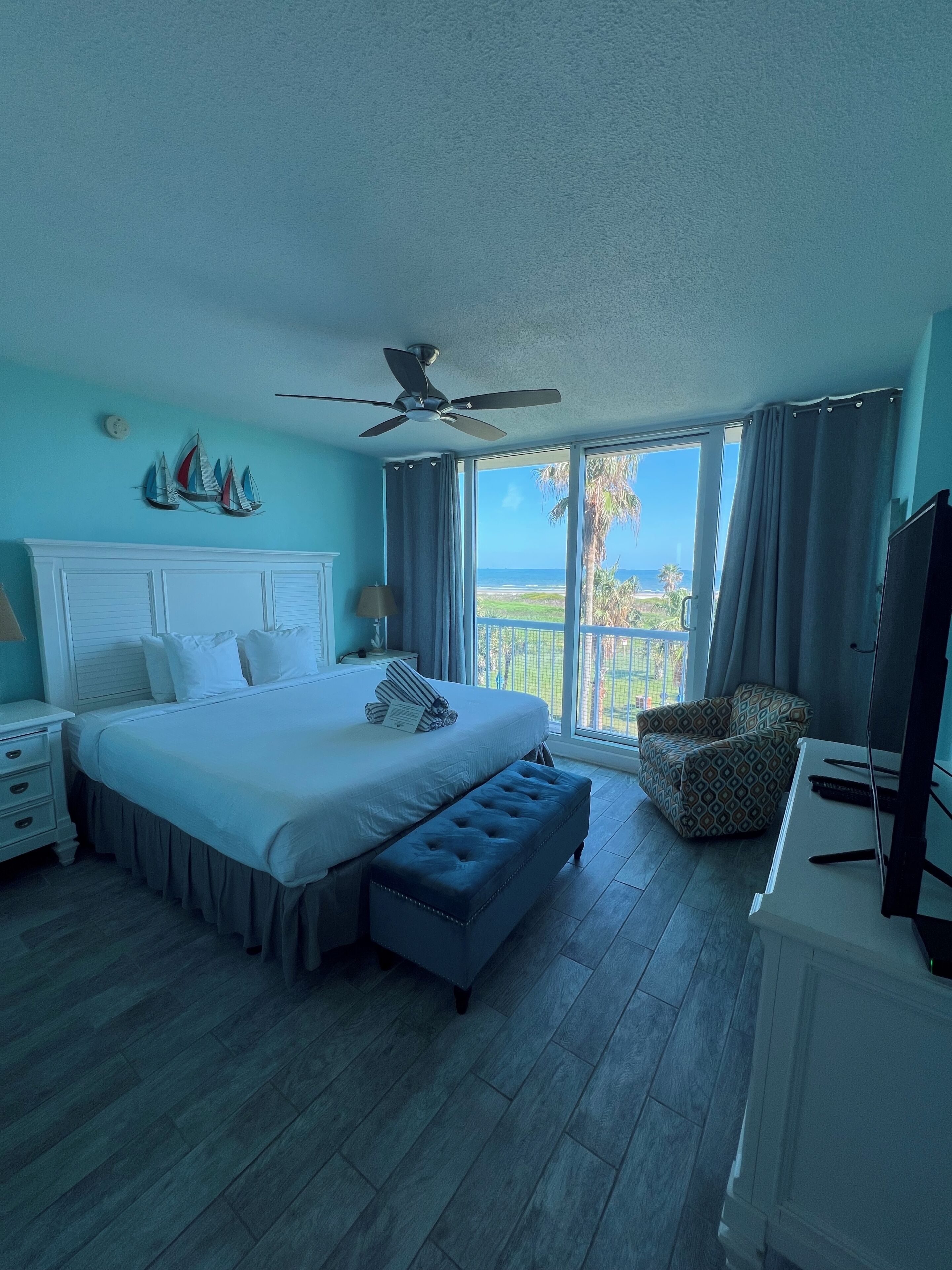 A bedroom with a bed, a ceiling fan, a desk with a computer, and a view of the ocean.