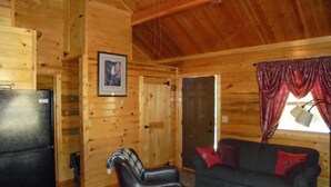A log cabin interior with a black refrigerator, a plaid chair, a dark blue couch with red cushions, and a patterned rug. - Come Stay in Belt Montana - Charming, Historic, Memorable (Belt)