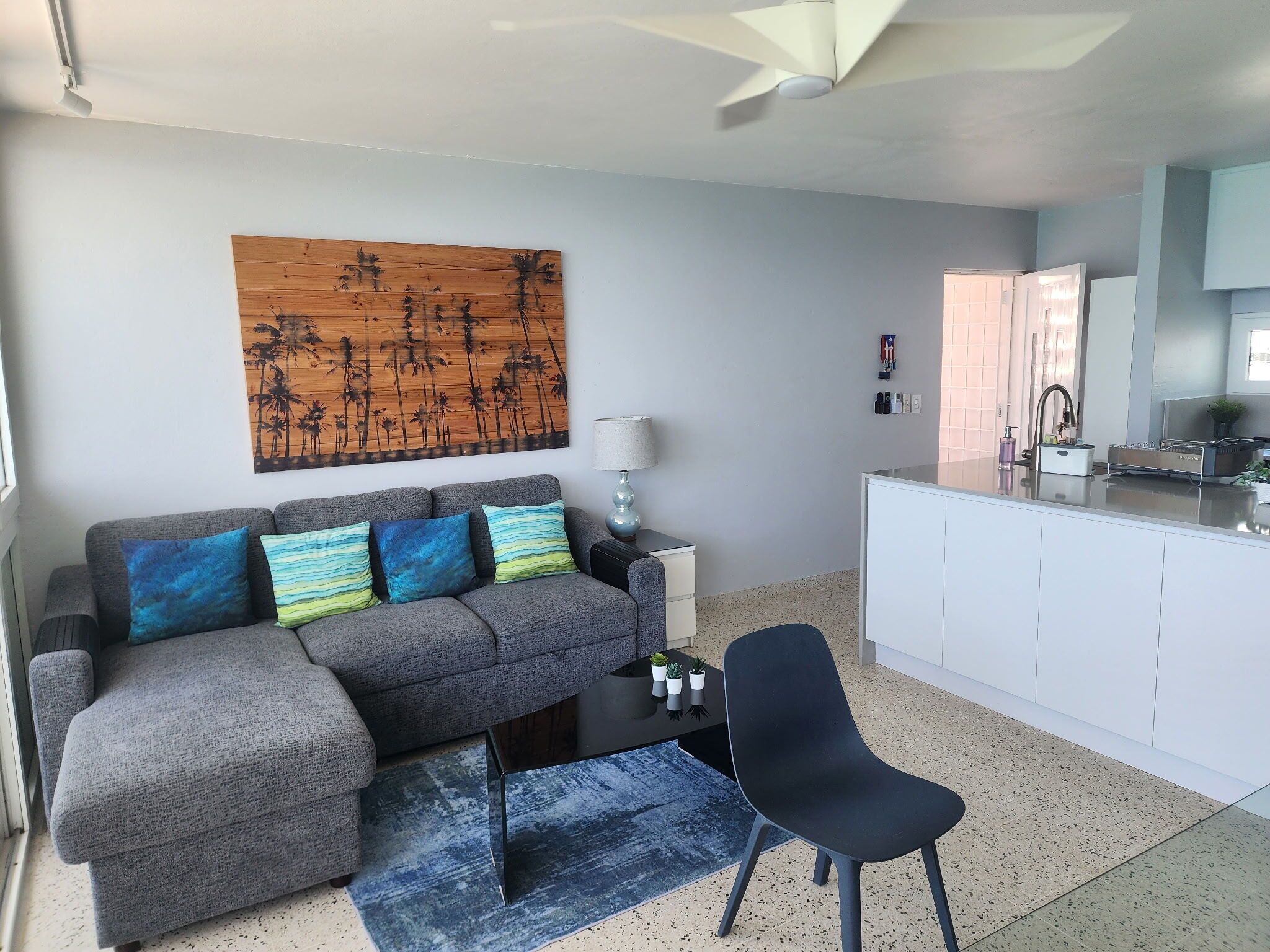 A modern living room with a grey sofa, a coffee table, a black chair, a wooden artwork, a lamp, and a kitchen area with a white cabinet and a sink.