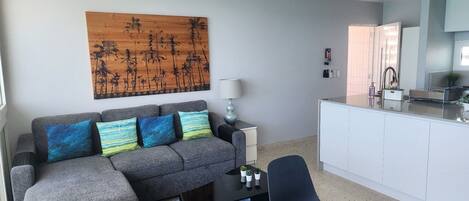 A modern living room with a grey sofa, a coffee table, a black chair, a wooden artwork, a lamp, and a kitchen area with a white cabinet and a sink.