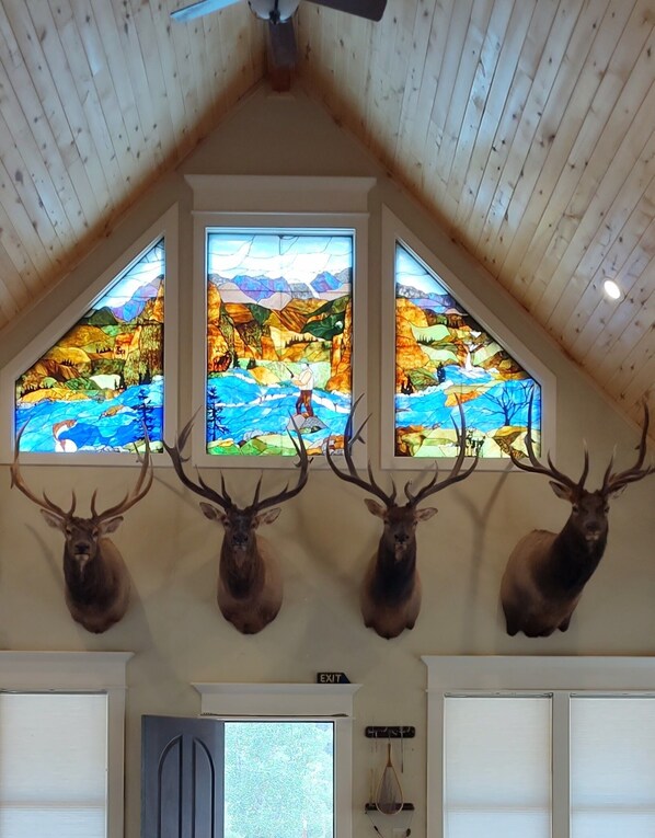 Une pièce dotée d’un plafond en bois, de quatre têtes de cerf fixées au mur et de trois vitraux représentant des paysages.