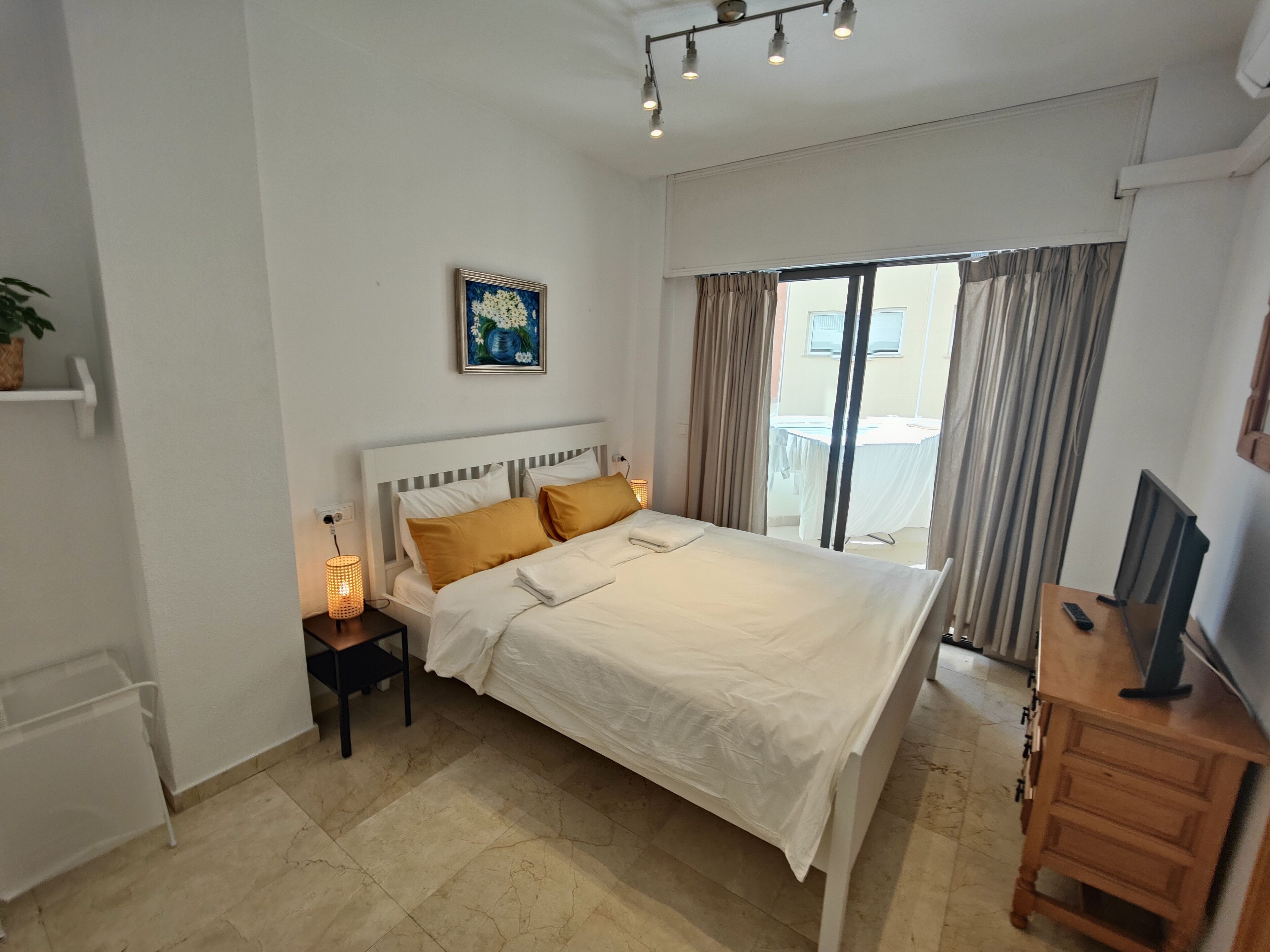A bedroom with a large bed, a bedside table with a lamp, a TV, and a window with curtains.