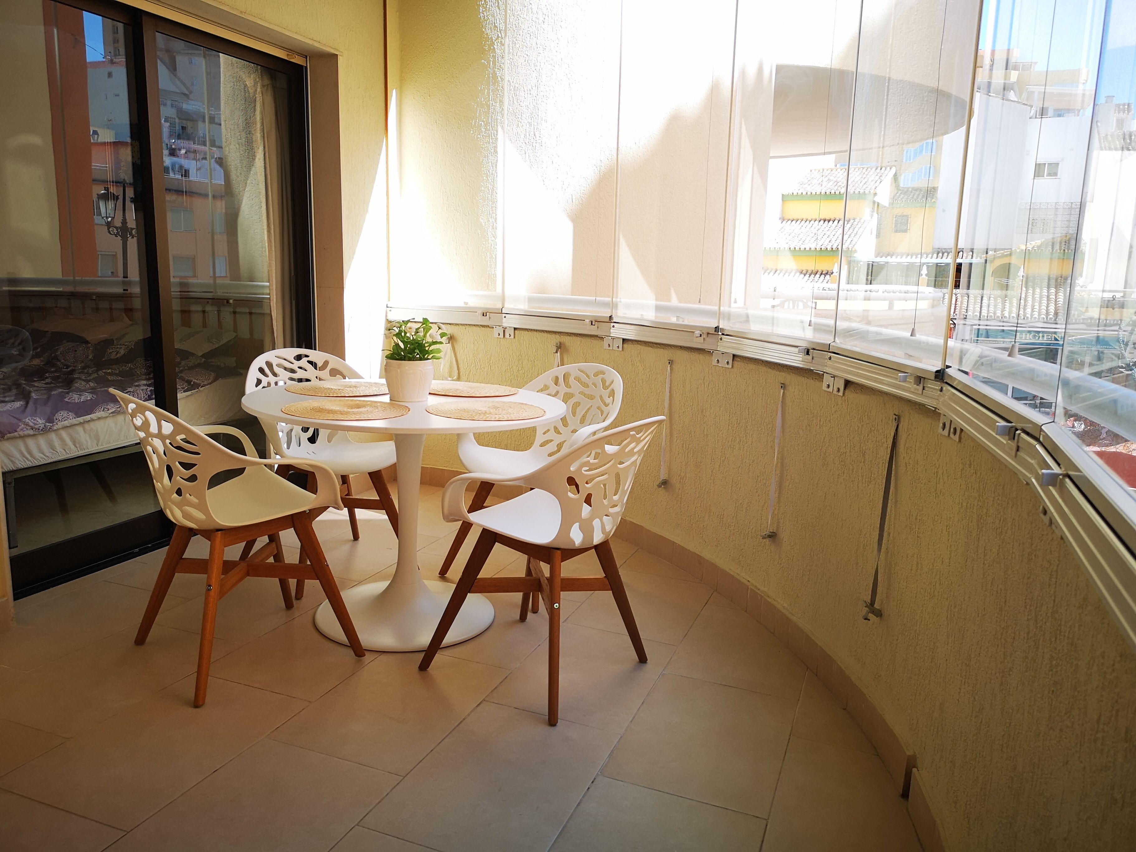 En balkon med et rundt bord og stole, en potteplante og udsigt til bygninger gennem en glasdør.