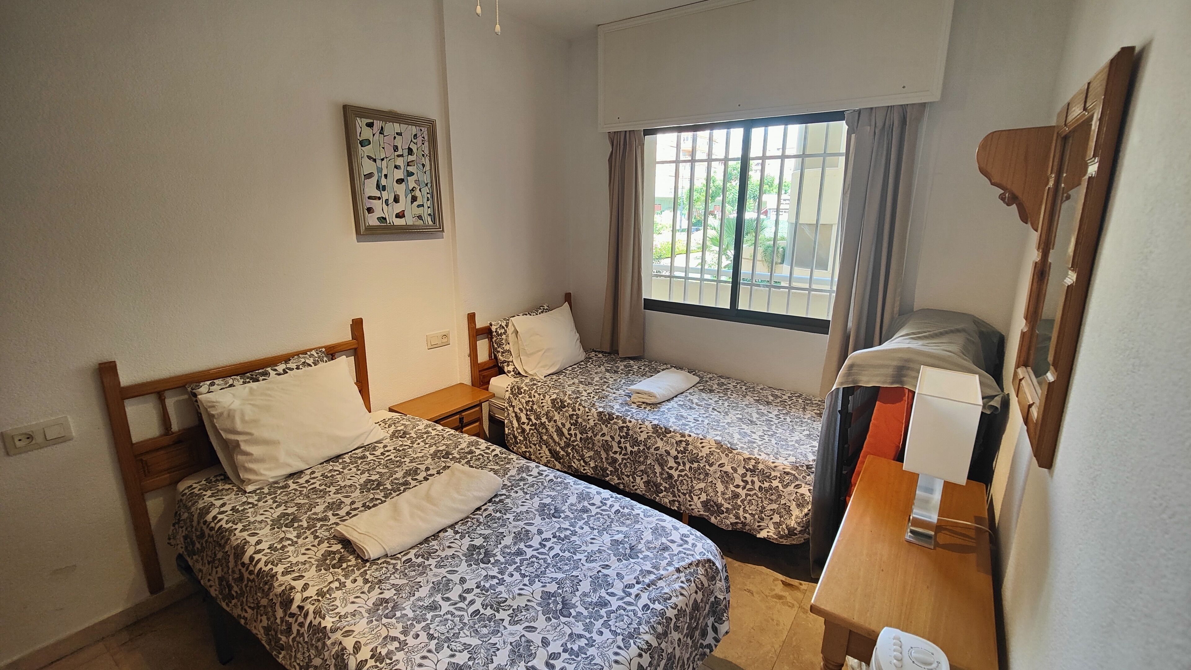 A small, clean room with two single beds, a wooden nightstand, a bedside table, a window with curtains, and a wall-mounted mirror.