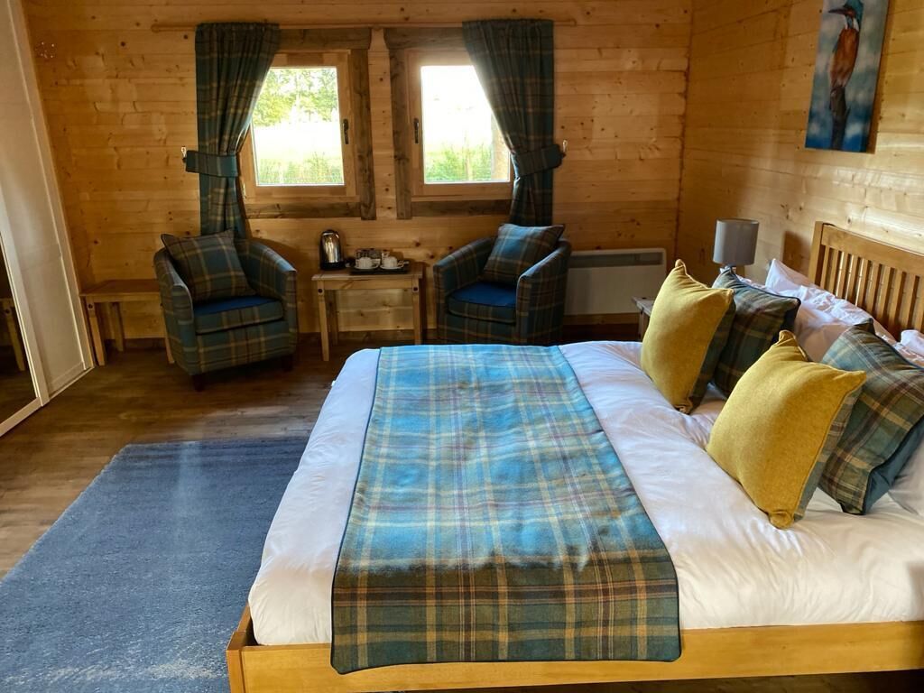 Un dormitorio de cabaña de madera con cama, dos sillones a cuadros, una mesita y una ventana con cortinas.