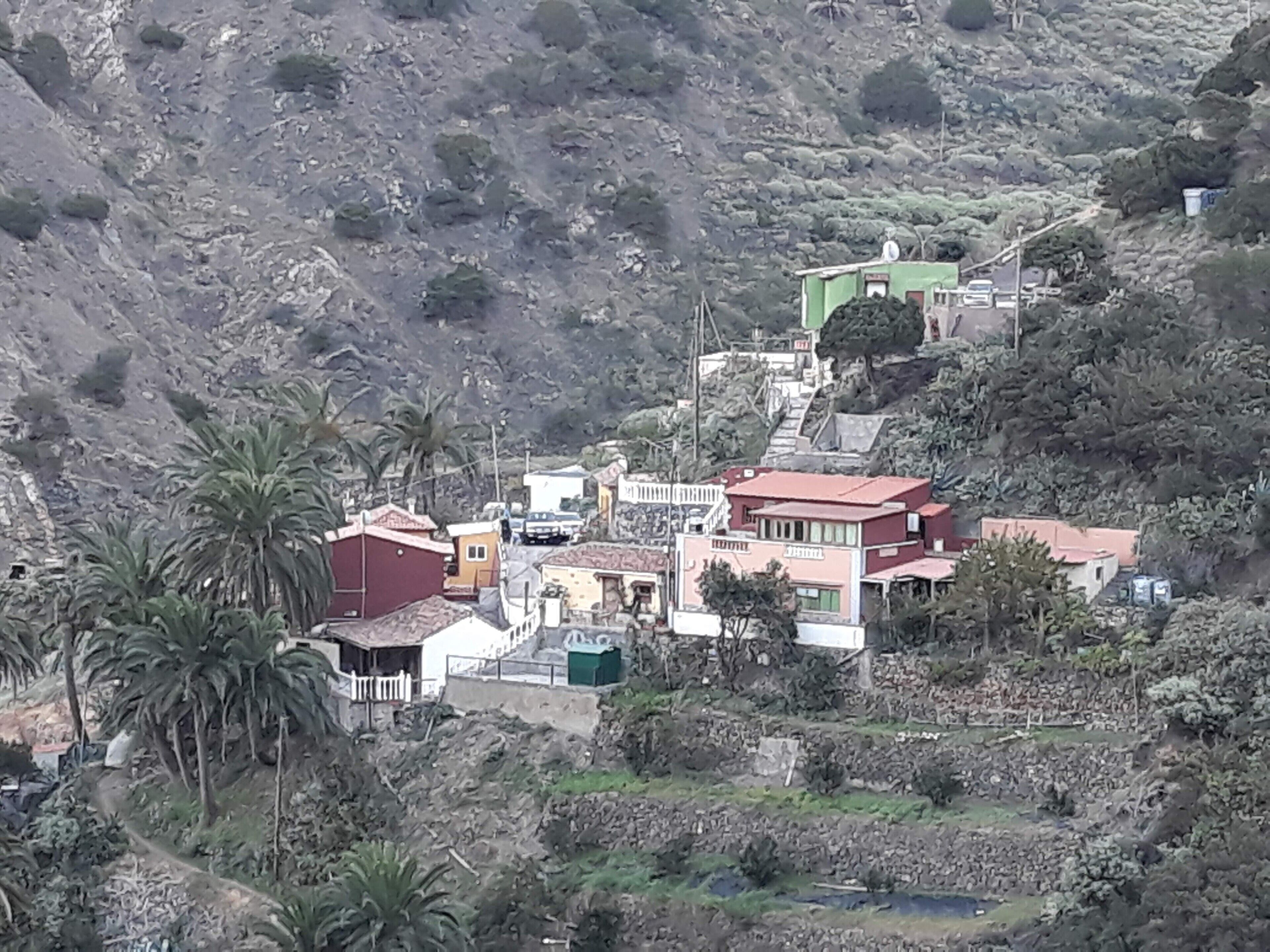 A hillside village with buildings, including a red-roofed house and a green building, surrounded by palm trees and rocky terrain.
